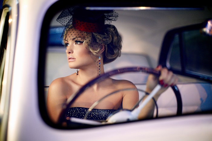 glamorous blonde woman with pink lipstick and vintage hat driving an old luxury car in evening light