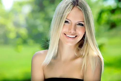 Smiling blonde woman outdoors surrounded by greenery, representing elegance, beauty, and a luxury lifestyle