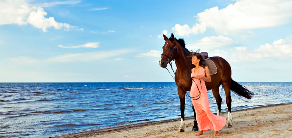 Elegant woman in a flowing pink dress walking with a horse along a sandy beach by the sea