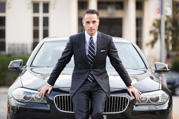 man in a suit sitting on the hood of a car with blurred cityscape in the background